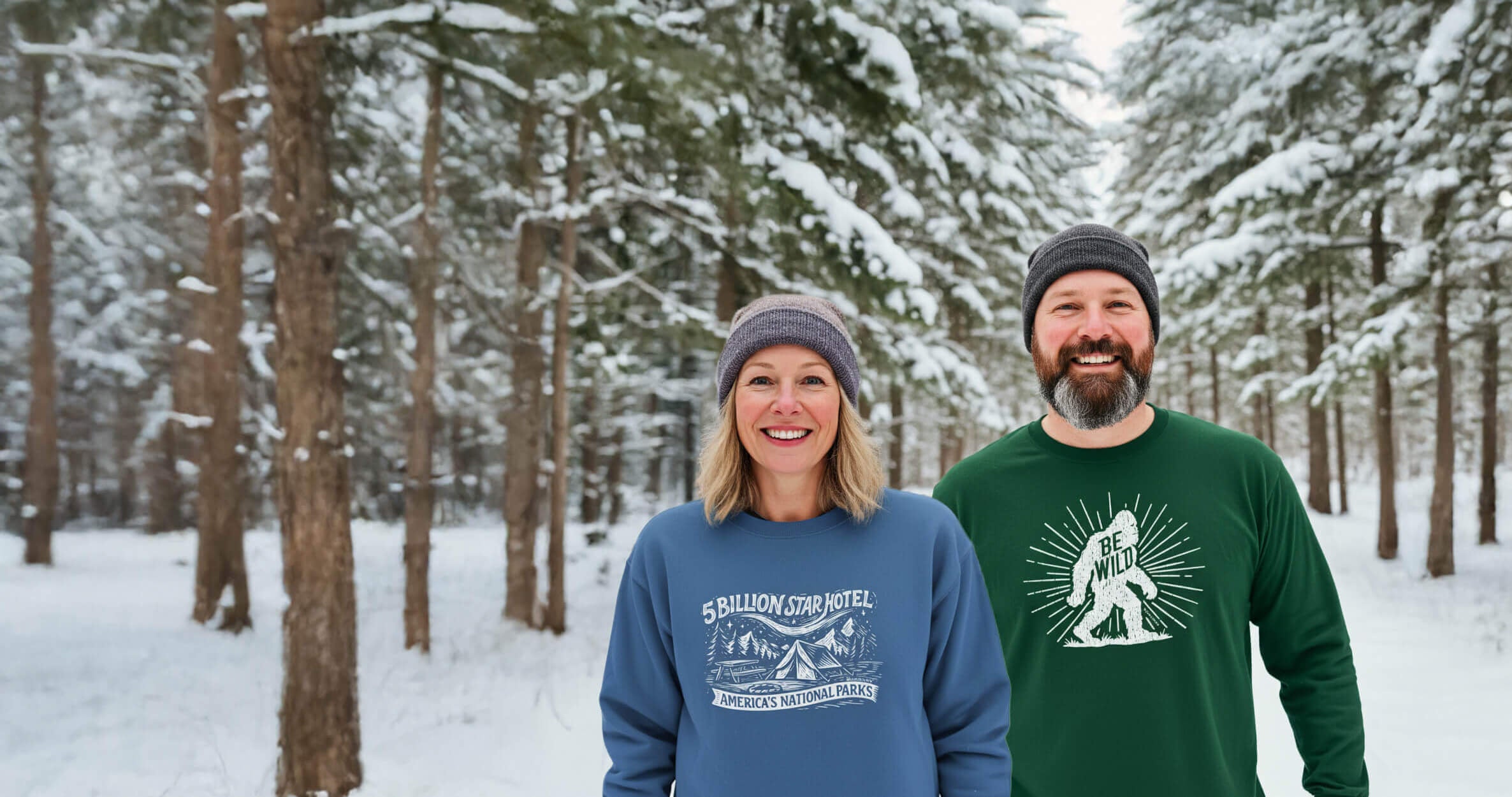 Two people standing in a snowy forest wearing sweatshirts with visible designs.